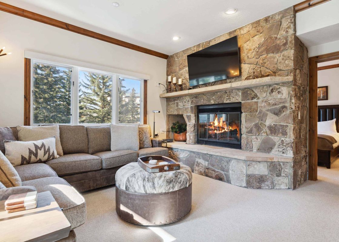 A cozy living room in The Charter C1205 with a stone fireplace, large sectional sofa, and windows overlooking pine trees.