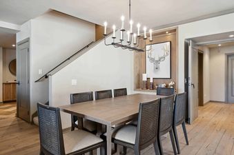 The dining area features a large wooden table and eight chairs with woven backs, lit by a stylish black chandelier, with a recessed wall niche displaying deer head artwork and decorative candles.