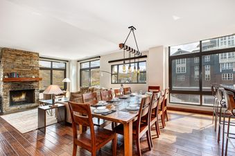 A spacious dining area with a long wooden table and chairs, set for a meal, transitions into a comfortable living room with a stone fireplace and large windows showcasing an outdoor view, all within the Lowell 402.