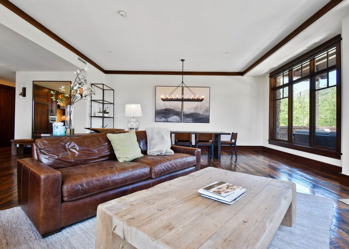 The comfortable living area of Arrowleaf Lodge 107 features a brown leather sofa, a wooden coffee table, and an adjacent dining area, with large windows offering a view of the outdoors.