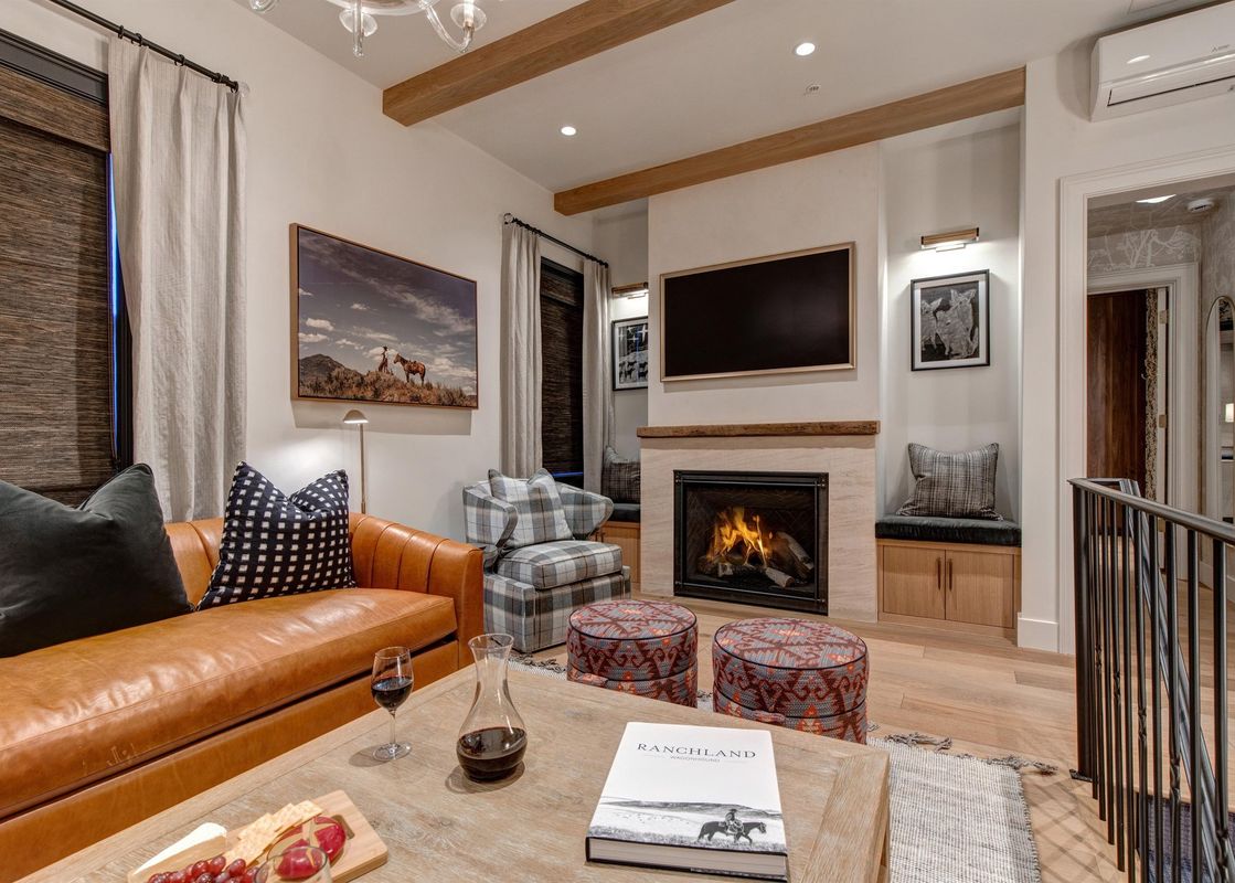 A living room with a fireplace, a large leather couch, a coffee table with a glass of wine and a book, and a large window with a view of the mountains.