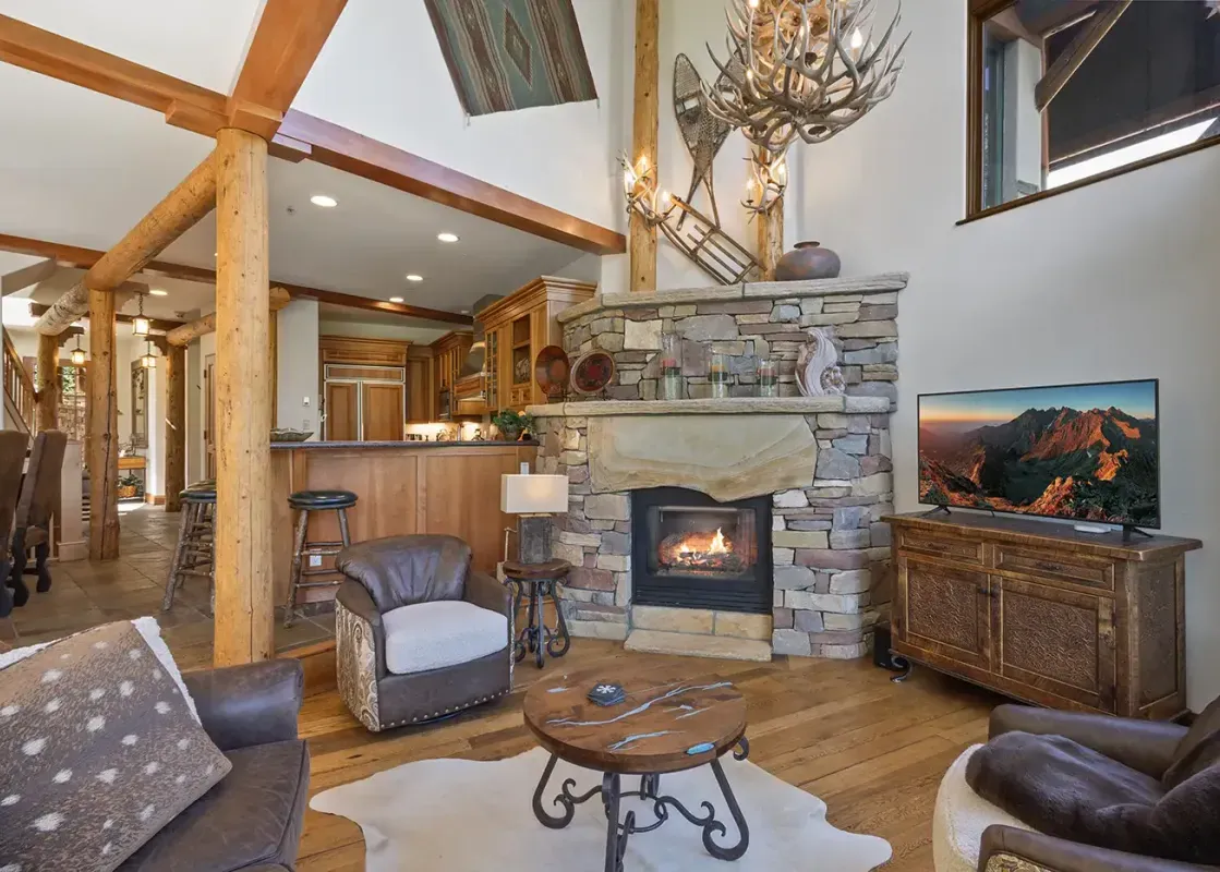 A cozy open-concept living area with a stone fireplace, rustic wooden beams, and an antler chandelier, featuring comfortable seating and a view into the adjacent kitchen and dining area.