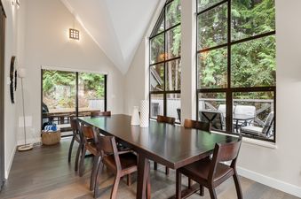 A spacious dining room at Whistler Pinnacle Ridge 42 with a dark wood table and chairs, large windows overlooking a lush forest, and a contemporary floor lamp.
