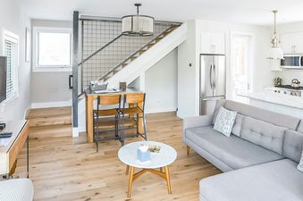 Living room with gray sectional sofa, coffee table, wood floors, kitchen counter and white cabinets in the background, and a wood staircase with an iron railing leading to the upper floor.