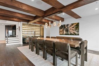 A sophisticated dining room at Stag Lodge 11 4BR features a long wooden table, modern pendant lighting, comfortable chairs, and a large built-in wine cellar, all under a ceiling with exposed wooden beams.