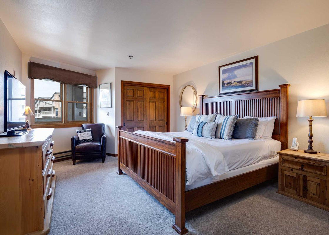 A spacious bedroom at Chateau Chamonix CX222 featuring a king-sized bed, wooden dresser with a TV, a cozy armchair, and a window with an exterior view.