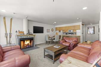 A cozy living room and kitchen area at Fashion Coalition 402, featuring red leather seating, a lit fireplace, a dining table, and a well-equipped kitchen with wooden cabinetry.