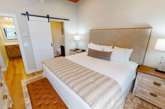 A cozy bedroom with a king-sized bed, a white duvet, and a patterned throw at the foot of the bed. There is a side table with a lamp, and a sliding barn door leading to a bathroom.