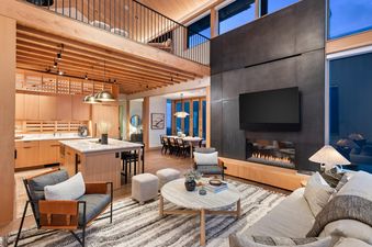 A spacious, modern living area with an open-concept kitchen, dining space, and a cozy lounge centered around a fireplace and television, featuring warm wood accents and abundant natural light.