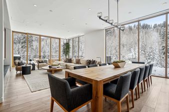 A wide shot of Founders Place 1701's open-concept dining and living area, featuring a long wooden dining table and a beige sectional sofa, all framed by large windows showcasing a snowy forest.