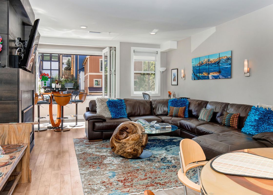 A modern living room featuring a brown leather sectional sofa with colorful pillows, a unique wooden coffee table, a breakfast bar, and large bi-fold doors opening to an exterior view.