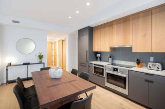 A modern kitchen and dining area with light wood and dark gray cabinetry, stainless steel appliances, a dark wood dining table, and a circular mirror above a console table.