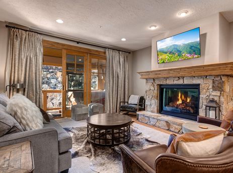 The warm and inviting living room of Town Pointe C304 features a stone fireplace, comfortable seating, and a TV, with large glass doors opening to a scenic balcony.