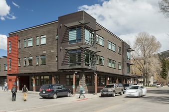 The Pied a Terre 202 building presents a modern facade with dark wood siding, red accents, and large windows, set in a lively urban environment with mountains and trees in the background.