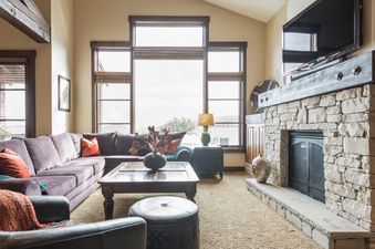 A cozy living room in Silver Star 3302 features a large purple sectional sofa, a stone fireplace with a television above it, and expansive windows offering scenic views.