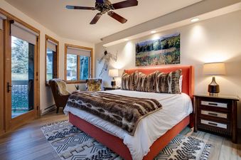 The bedroom at Chateau Chamonix CX322 features a large bed with a vibrant orange headboard, faux fur bedding, two nightstands with lamps, a cozy armchair, and large windows offering an outdoor view.
