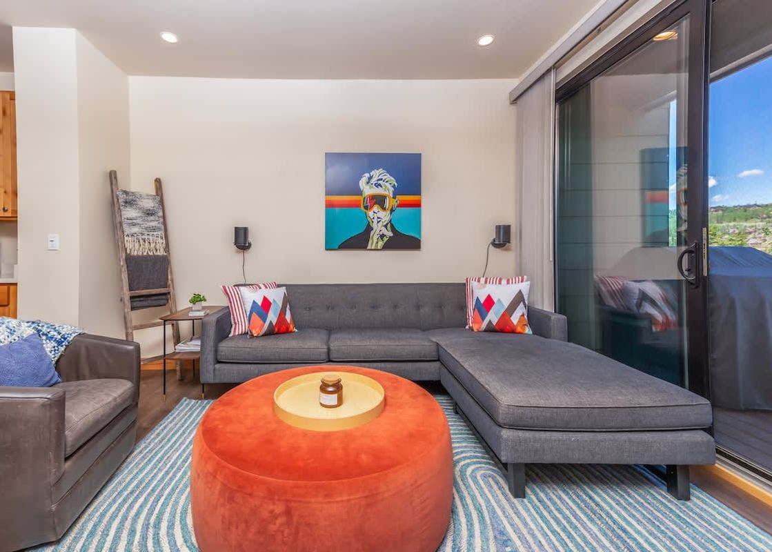 The living room at Basecamp Luxe features a gray sectional sofa with colorful throw pillows, an orange ottoman, and a modern art piece on the wall.