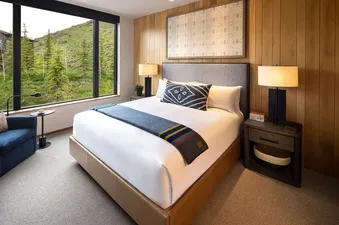 A king-sized bed with white linens and a blue, gold, and brown throw blanket is flanked by two nightstands with lamps, all set against a wood-paneled wall, while a large window offers a view of a lush green mountain landscape.