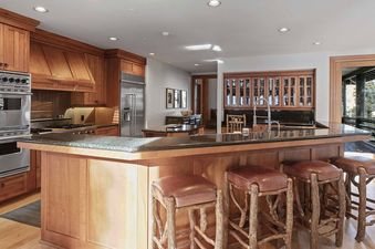 A luxury kitchen with warm wood cabinetry, stainless steel appliances, a large granite island with a sink, and three rustic wooden bar stools with leather seats.