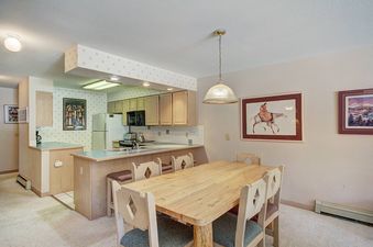 Dining room with a large wooden table, six chairs, and a kitchen in the background.