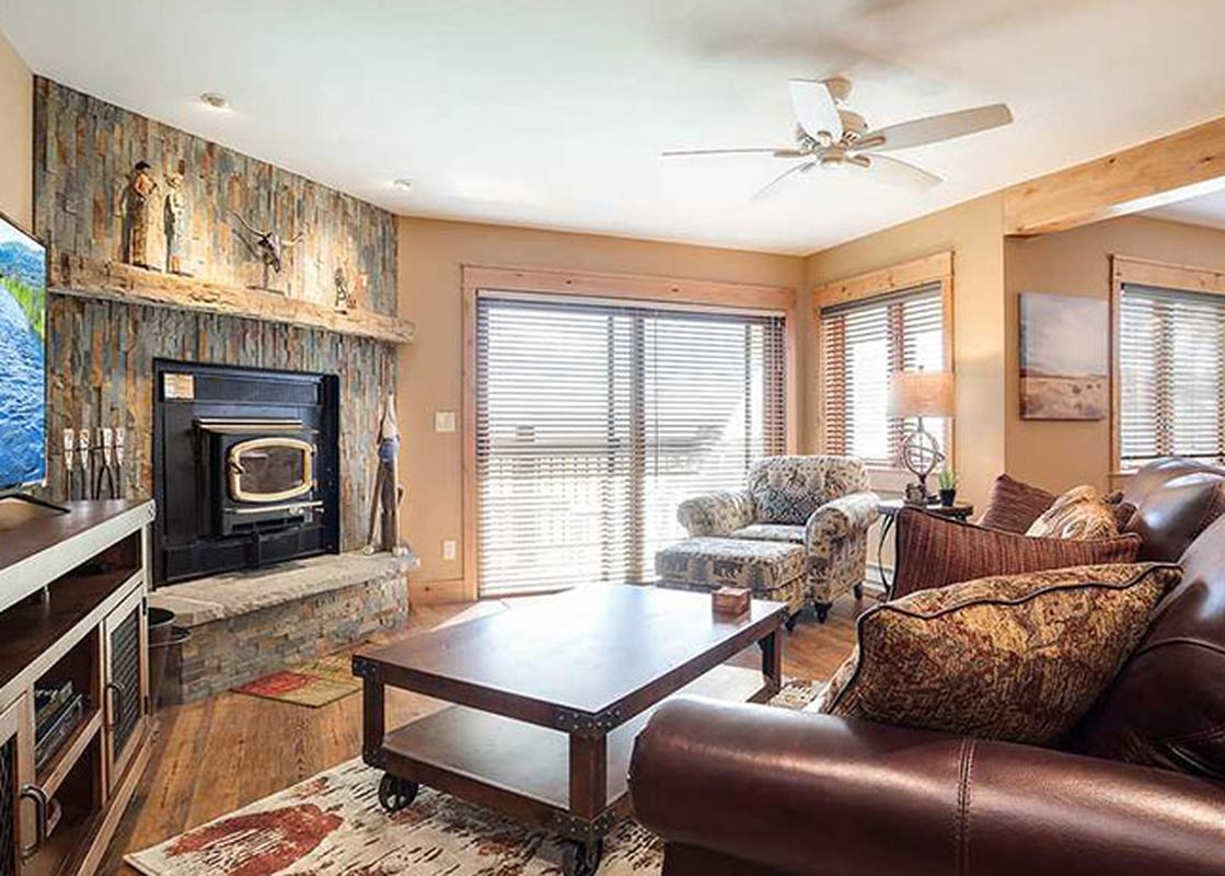 A rustic-style living room with a stone fireplace, brown leather sofa, wooden coffee table, and a TV showing a river scene.