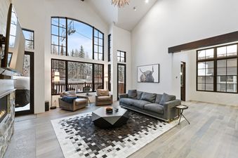 A spacious living room at Slopeside 129 with large windows showcasing a snowy forest and ski lifts, furnished with a gray sofa, leather armchairs, and a geometric coffee table on a patterned rug.