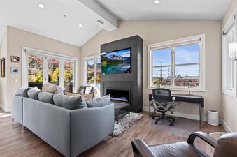 A modern living room in Rio Grande 301 with a grey sofa, a fireplace with a TV, a desk with a chair, and large windows showcasing an autumn landscape.