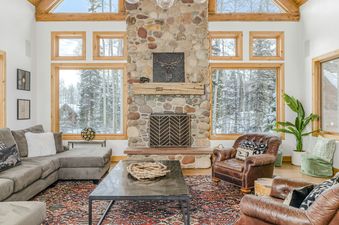 A cozy great room at Palmyra 94 Mountain Tranquility featuring a stone fireplace, plush seating, and expansive windows with views of a snowy forest.
