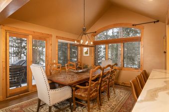 The dining room at Fairview Lodge features a large wooden dining table with various chairs and a view of the snowy outdoors through a large window and glass doors.