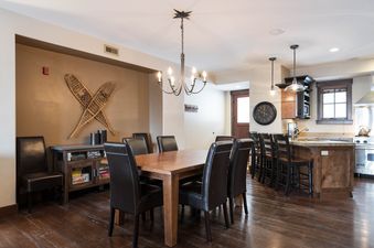A dining table with six chairs and an adjacent kitchen with an island, bar stools, and stainless steel appliances are shown.