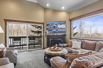 A living room with a leather sectional sofa, a coffee table, and a fireplace. The room has large windows with views of the mountains and trees.