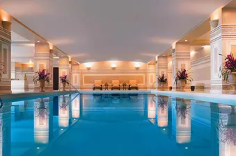 An indoor swimming pool with decorative pillars and lounge chairs. The pool has clear blue water with reflections of the pillars and ceiling.