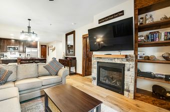 The open-concept living area at BlueSky Breckenridge 407 offers a seamless flow between the living room and kitchen, featuring a stone fireplace, large TV, comfortable sectional, and a well-equipped kitchen with dark wood cabinetry.