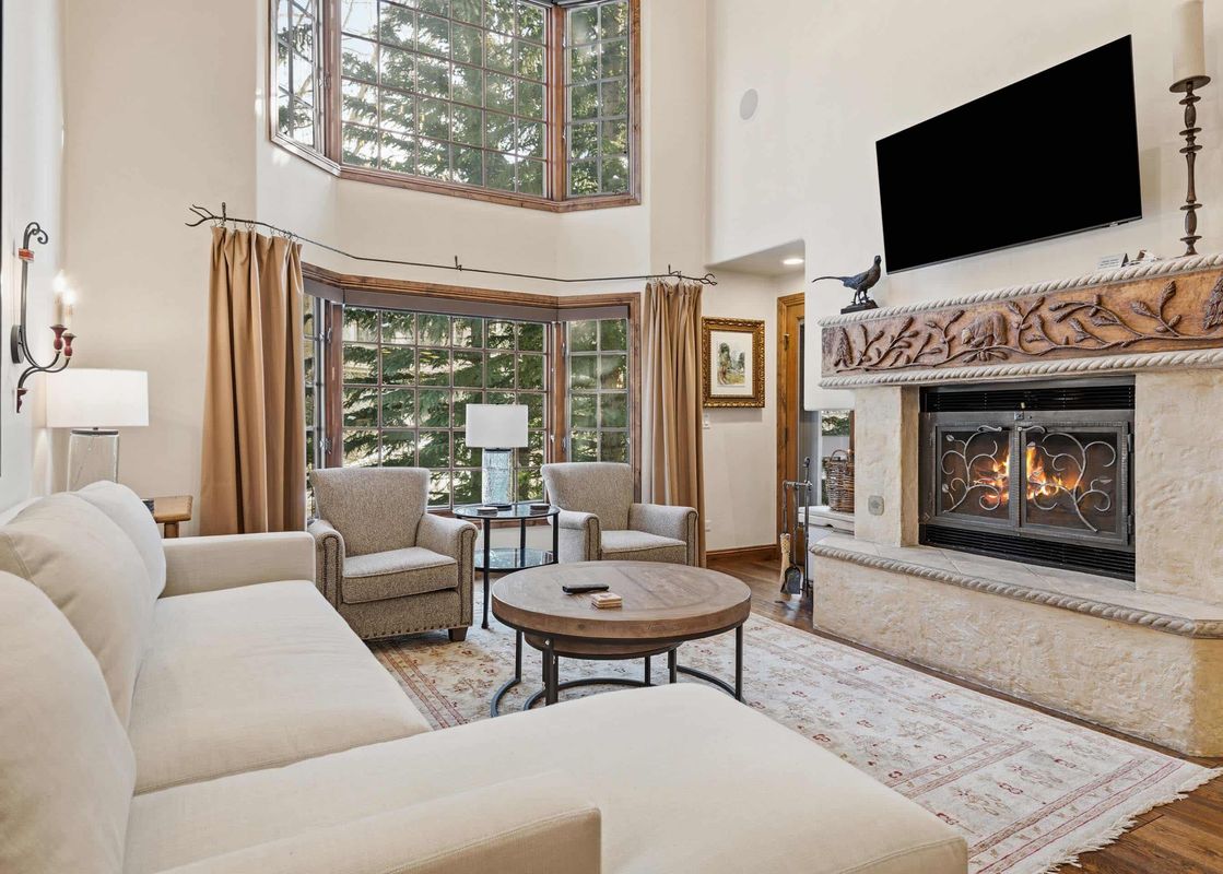 A spacious and inviting living room in The Charter G230 features a comfortable sectional sofa, two accent chairs, a stone fireplace, and large windows with outdoor views.