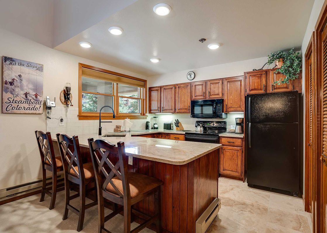 A fully equipped kitchen with wooden cabinets, black appliances, a central island with bar seating, and a large window providing natural light in Chateau Chamonix CX233.