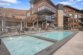 An outdoor pool and hot tub area with lounge chairs and umbrellas, set against a modern building with wood siding and balconies, and a mountain in the background.