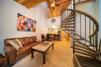 The living area of Creekside 403 features a brown leather sectional sofa, a coffee table, and a colorful bear painting on the wall. A spiral staircase leads upstairs.