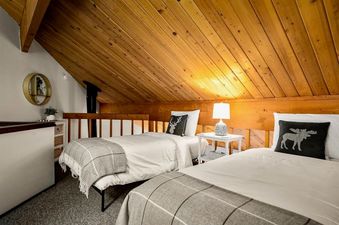 A cozy bedroom at Cedar Creek Cabin featuring two twin beds with white bedding and grey plaid throws, rustic wooden ceilings, and a decorative nightstand with a lamp.