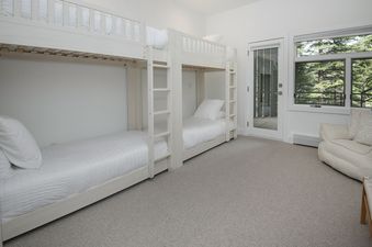 A bright bunk room in Rockledge 325 with two sets of white bunk beds, a large window revealing evergreen trees, and a cozy armchair.