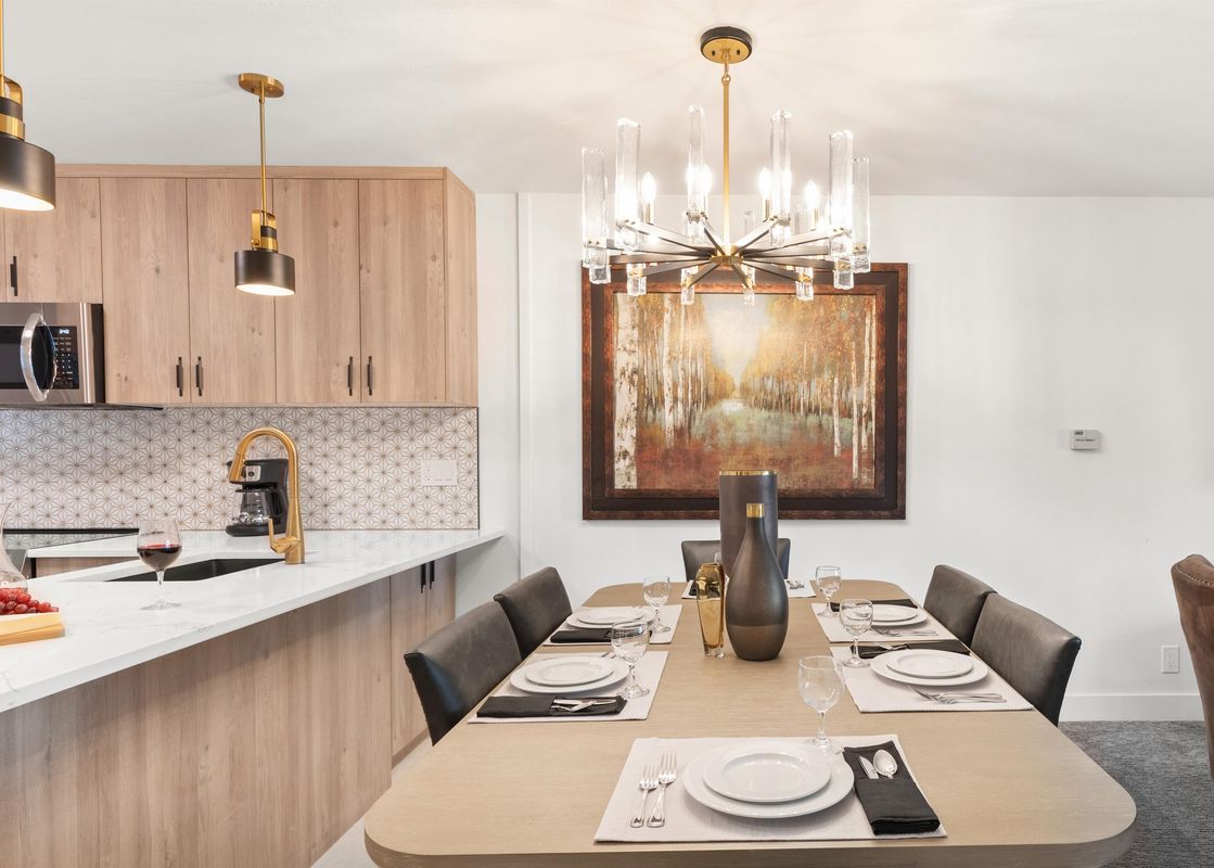 A modern dining room and kitchen with a wooden dining table set with white plates, dark gray napkins, and glassware, featuring a golden and black chandelier above and a nature-inspired painting on the wall, with a light wood kitchen featuring white counte