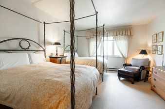 A cozy bedroom in Strawberry Park 603 features two queen-sized beds with white linens, soft beige comforters, and ornate metal frames, alongside a comfortable armchair, a wooden dresser, and ambient lighting from lamps and a large window.
