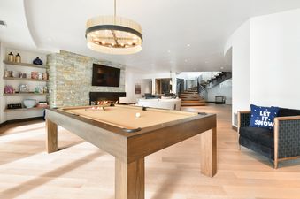 A spacious game room at Iron Mountain Chalet with a pool table, stone fireplace, flat-screen TV, and decorative shelves, leading to a stylish staircase.
