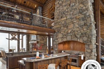 A rustic-style kitchen with a large stone fireplace, a stainless steel range, a wooden island with bar stools, and a loft area above, set against a backdrop of a snowy outdoor view.