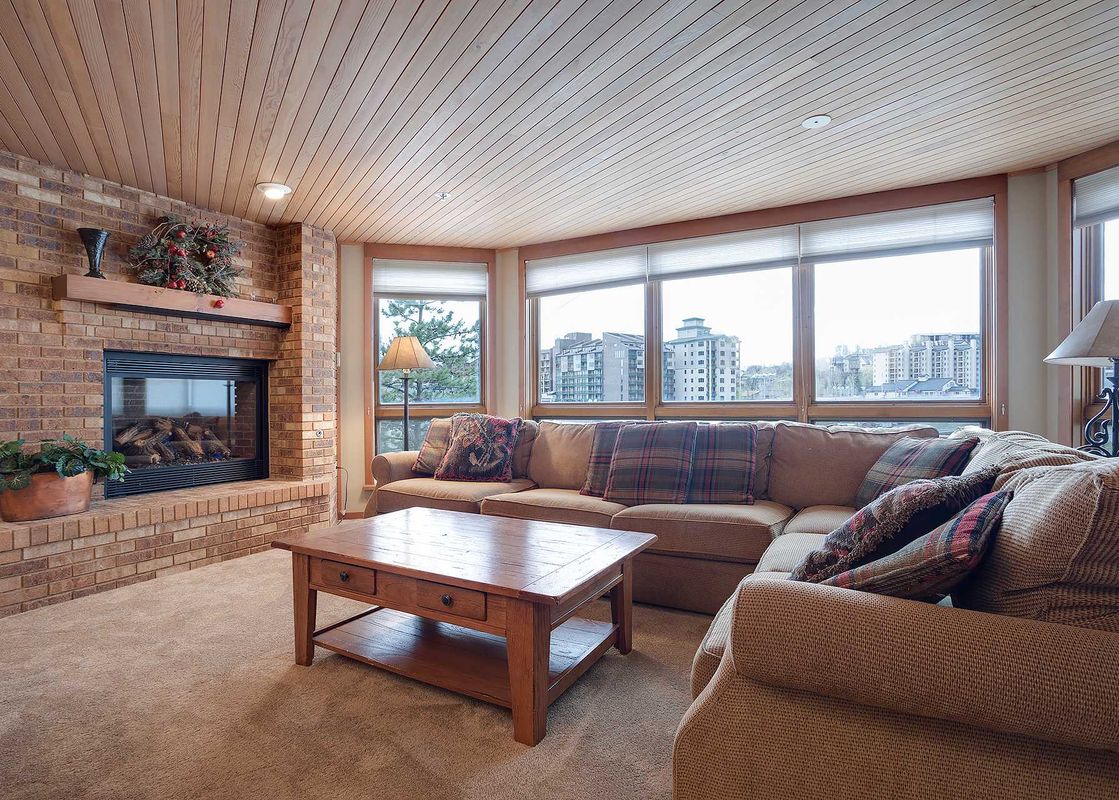 A cozy living room at Chateau Chamonix CX124 featuring a brick fireplace, a large sectional sofa, and a wooden coffee table, with windows offering city views.
