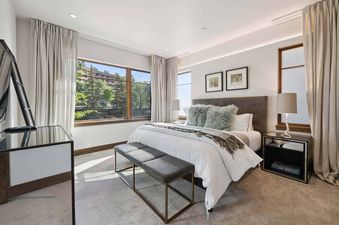 A bright and airy master bedroom with a king-sized bed, mirrored nightstands, and large windows offering a view of greenery.
