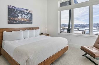 Modern bedroom with a wooden king-size bed, tan leather chair, and large windows looking out on a snowy landscape in the SkyRidge 1152 unit.