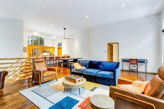 A spacious, open-concept living area with a blue velvet sofa, tan leather armchairs, a foosball table on a round coffee table, and an open kitchen with wooden cabinets and stainless steel appliances in the background.