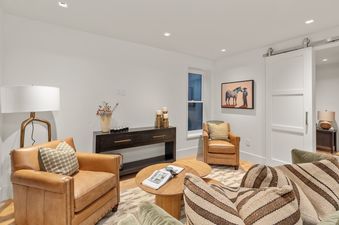 A cozy living room at the Depot House features two tan leather armchairs, a green sofa with striped pillows, a dark console table, and a painting of a cowboy and horse, all under soft lighting.