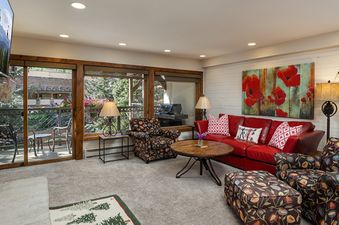 The Montaneros 211 living area, featuring a red leather sofa, two patterned armchairs, a wooden coffee table, and large windows opening to a balcony with a BBQ grill.