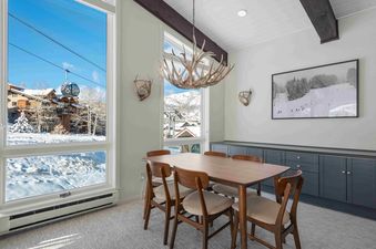 A dining room at Enclave 313 featuring a wooden table and chairs, an antler chandelier, and large windows revealing a snowy mountain landscape with a gondola.